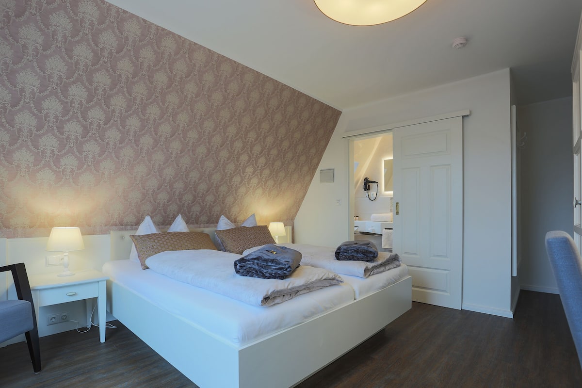 Quarto de hotel moderno com uma cama grande, mesas de cabeceira com abajures e um papel de parede estampado.