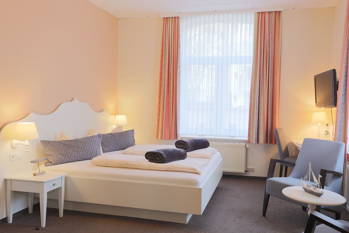 Quarto de hotel com cama, duas mesas de cabeceira, uma cadeira, televisão e janela com cortinas.