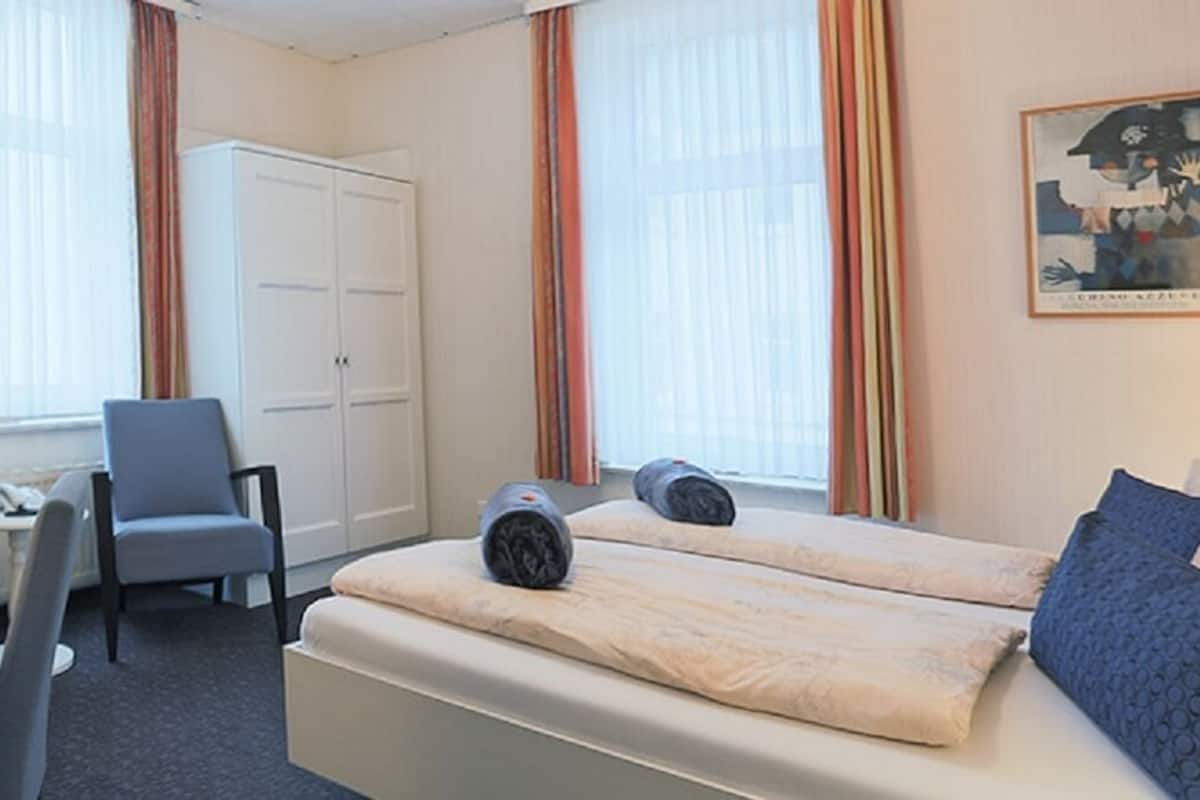 Quarto de hotel com cama, duas cadeiras, escrivaninha, televisão e guarda-roupa.