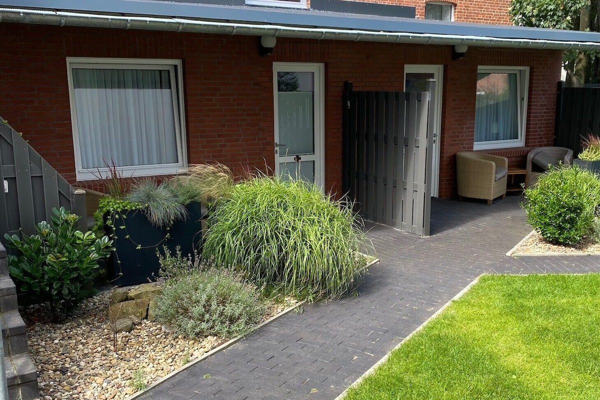 Um imóvel residencial com um caminho de entrada pavimentado que leva a porta da frente, uma pequena área de jardim com diversas plantas e um gramado bem cuidado.