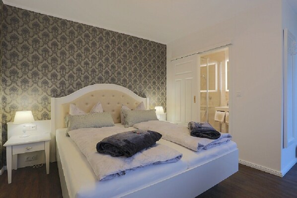 A neatly made bed with pillows and a blanket, a bedside table with a lamp, and a patterned wallpaper on the wall.