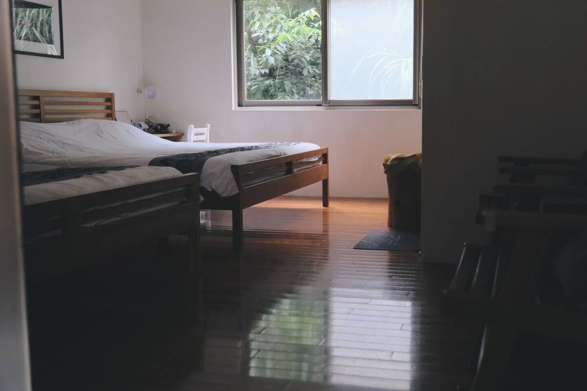 Une pièce avec un lit, une chaise, une fenêtre et une poubelle.