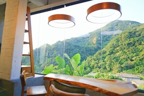 Une pièce avec une table et des chaises en bois, une fenêtre donnant sur une montagne et la verdure, et deux lampes suspendues.