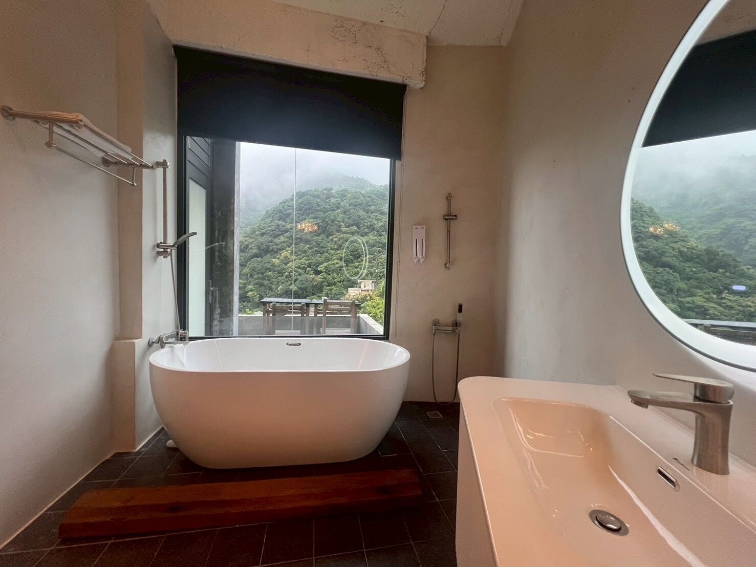 Un baño moderno con una bañera independiente, un espejo grande y vistas a un paisaje verde exuberante a través de un ventanal.