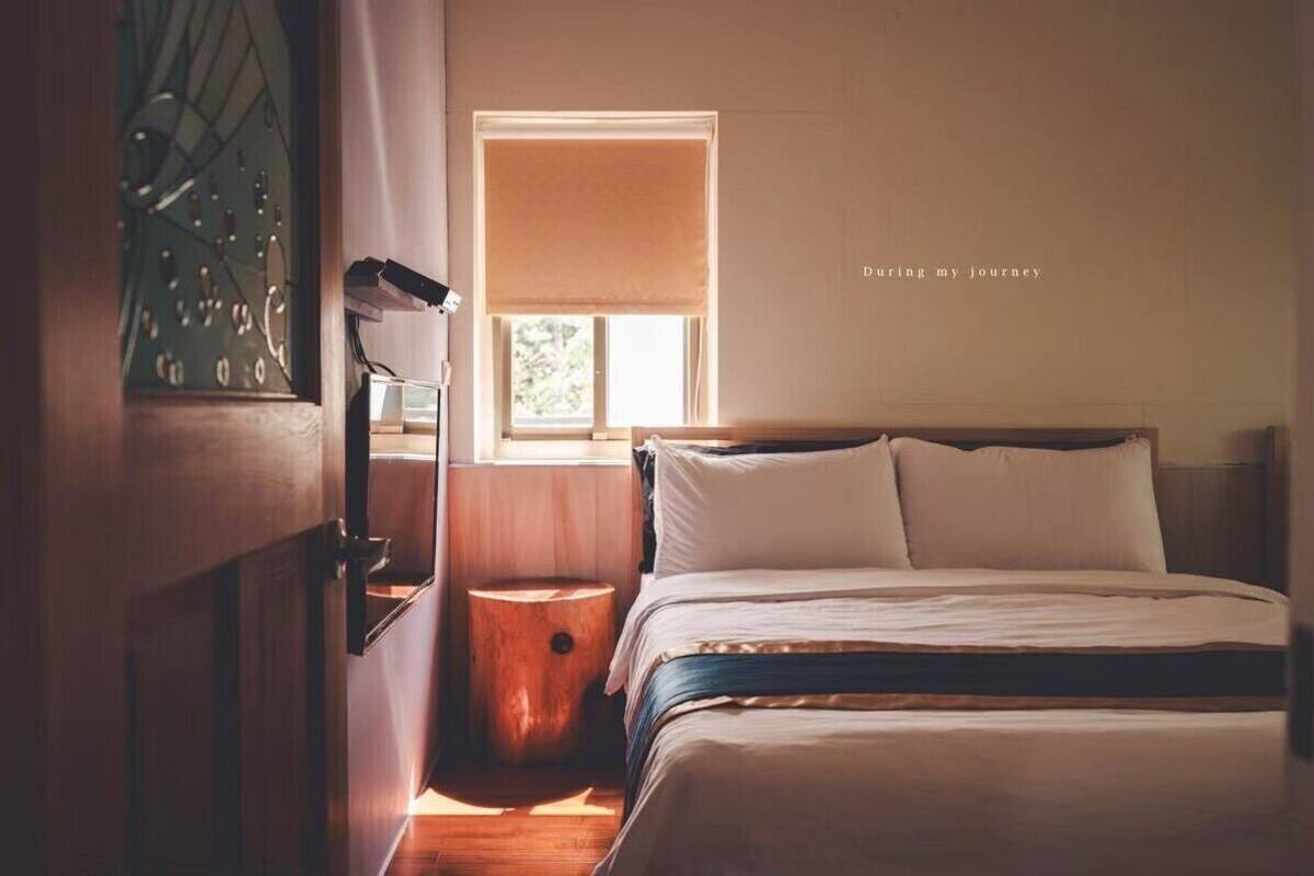 Une chambre à coucher avec un lit, une fenêtre avec un store, un tabouret en bois et une horloge murale.