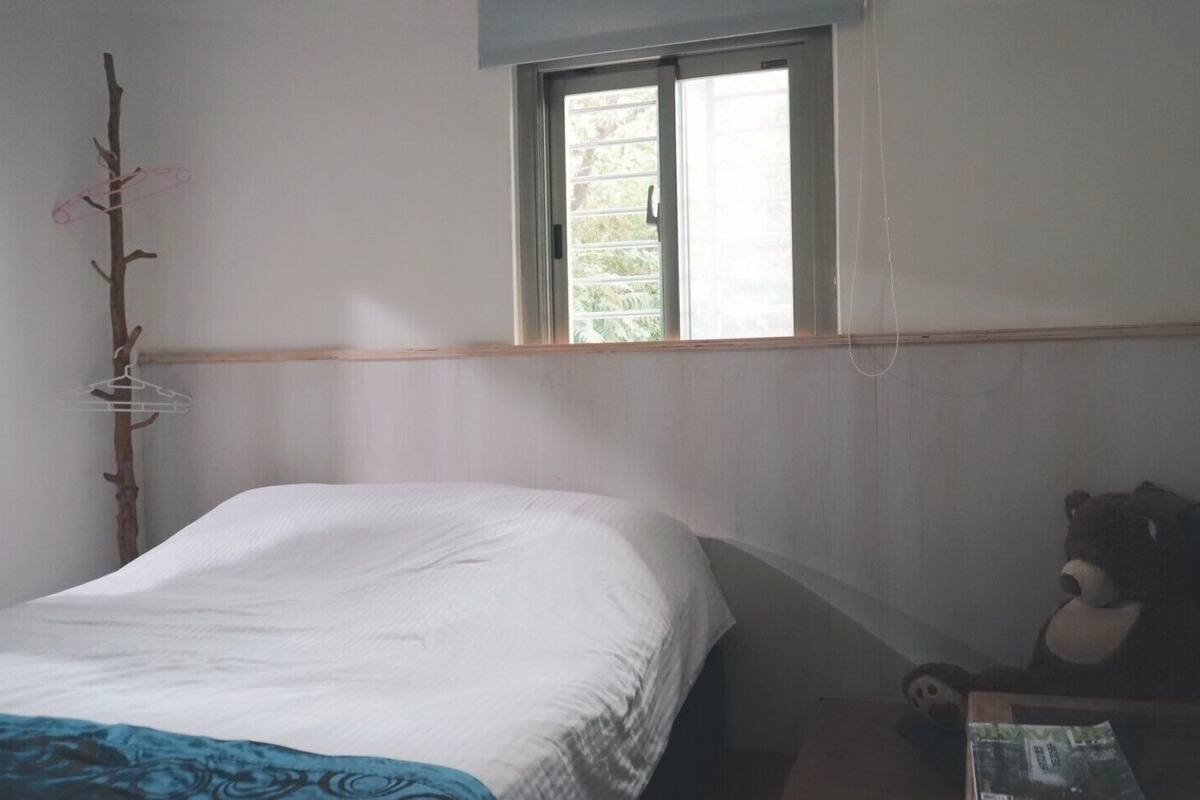 Une pièce simple, avec un lit, une chaise, une fenêtre et une étagère fixée au mur.