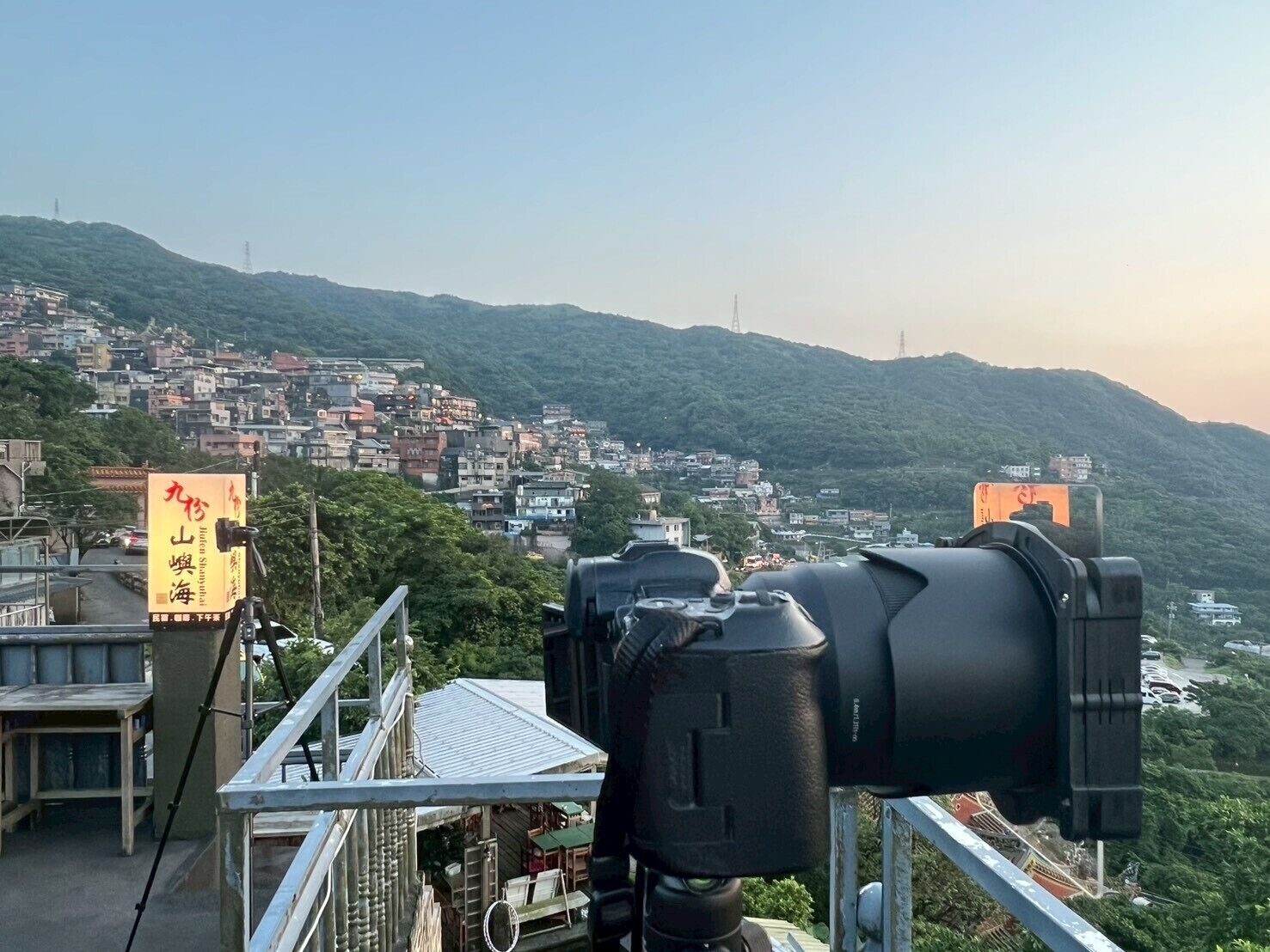 A camera on a balcony overlooking a densely packed hillside town with visible buildings and infrastructure.