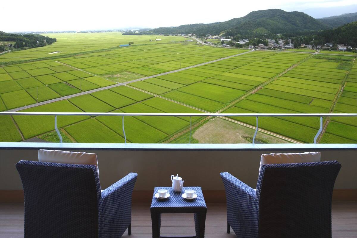 Ein Balkon mit Korbstühlen und einem kleinen Tisch, mit Blick auf ein weitläufiges Grünfeld mit einem Berg im Hintergrund.