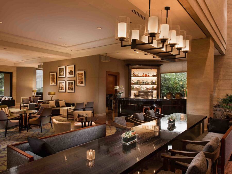 A spacious hotel lobby with a long dining table, comfortable seating, a bar area, and framed artwork on the walls.