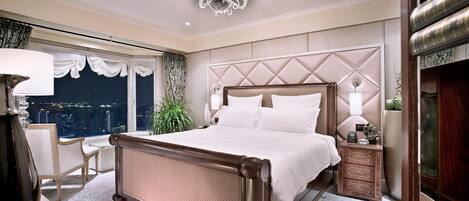 A spacious bedroom with a large bed, a nightstand, a chair, and a view of the city at night.
