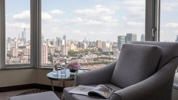 A room with a large window, a grey armchair, a small round table with a vase and glasses, and a magazine.
