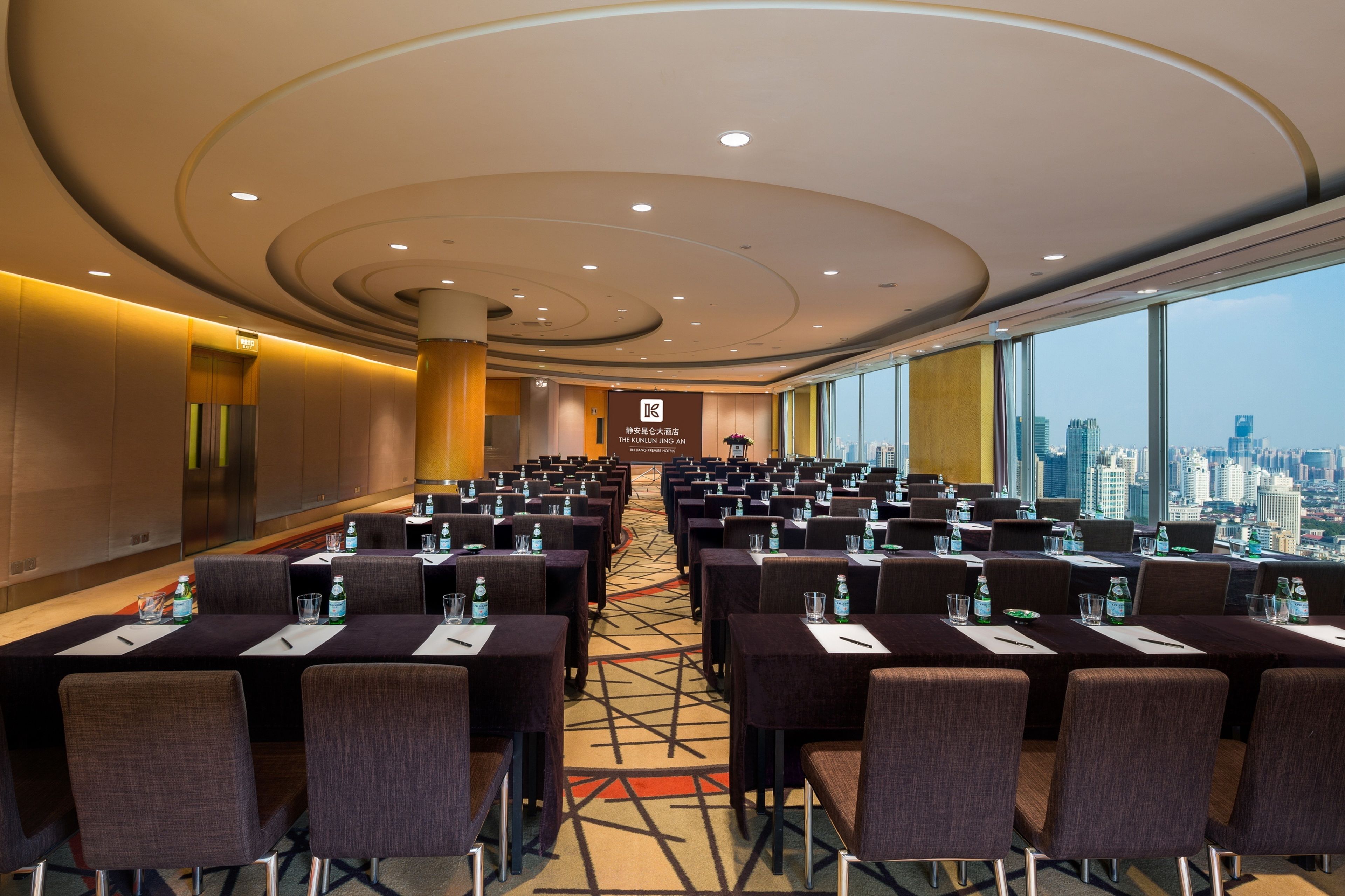 A conference room with rows of tables and chairs, a large window with a city view, and a modern ceiling design.