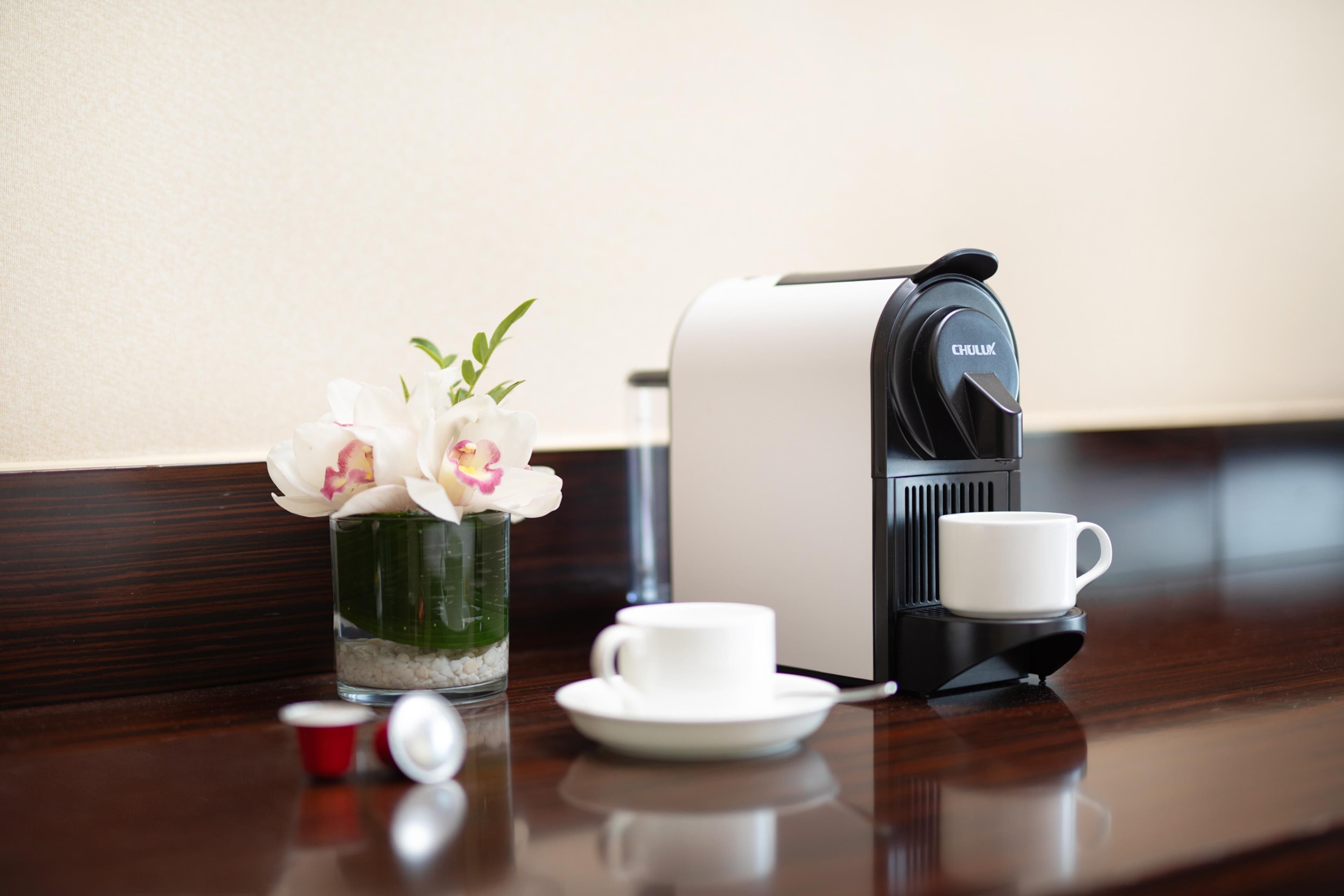 A coffee maker, a cup, and a vase with flowers on a wooden table.