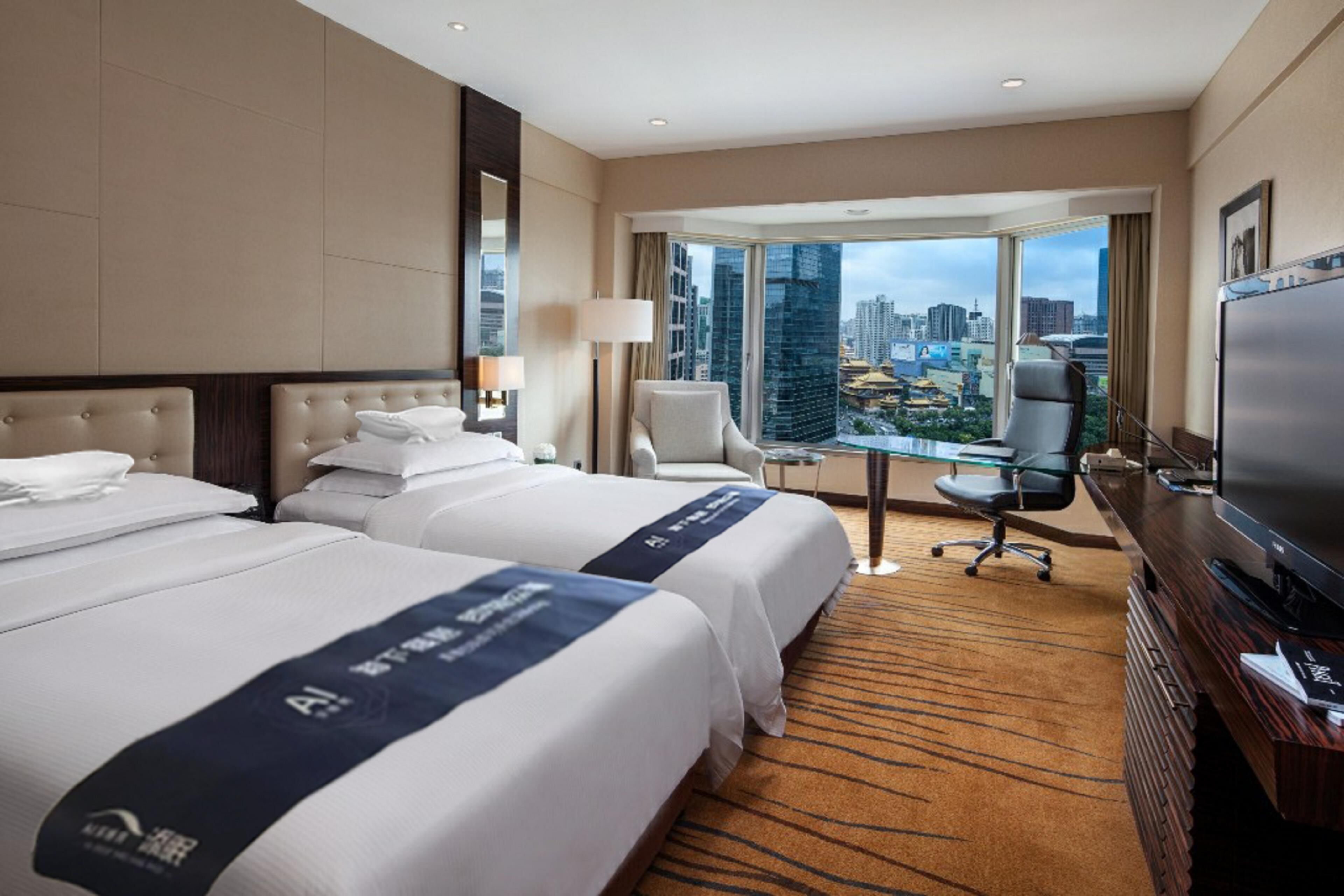 A hotel room with two beds, a desk, a chair, a television, and a large window offering a city view.