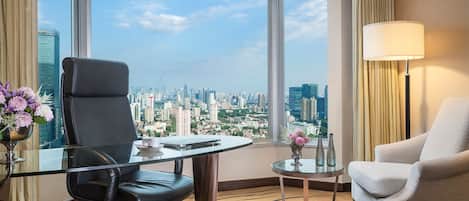 A hotel room with a city view, a glass table, a black office chair, a white armchair, and a small side table with a lamp.