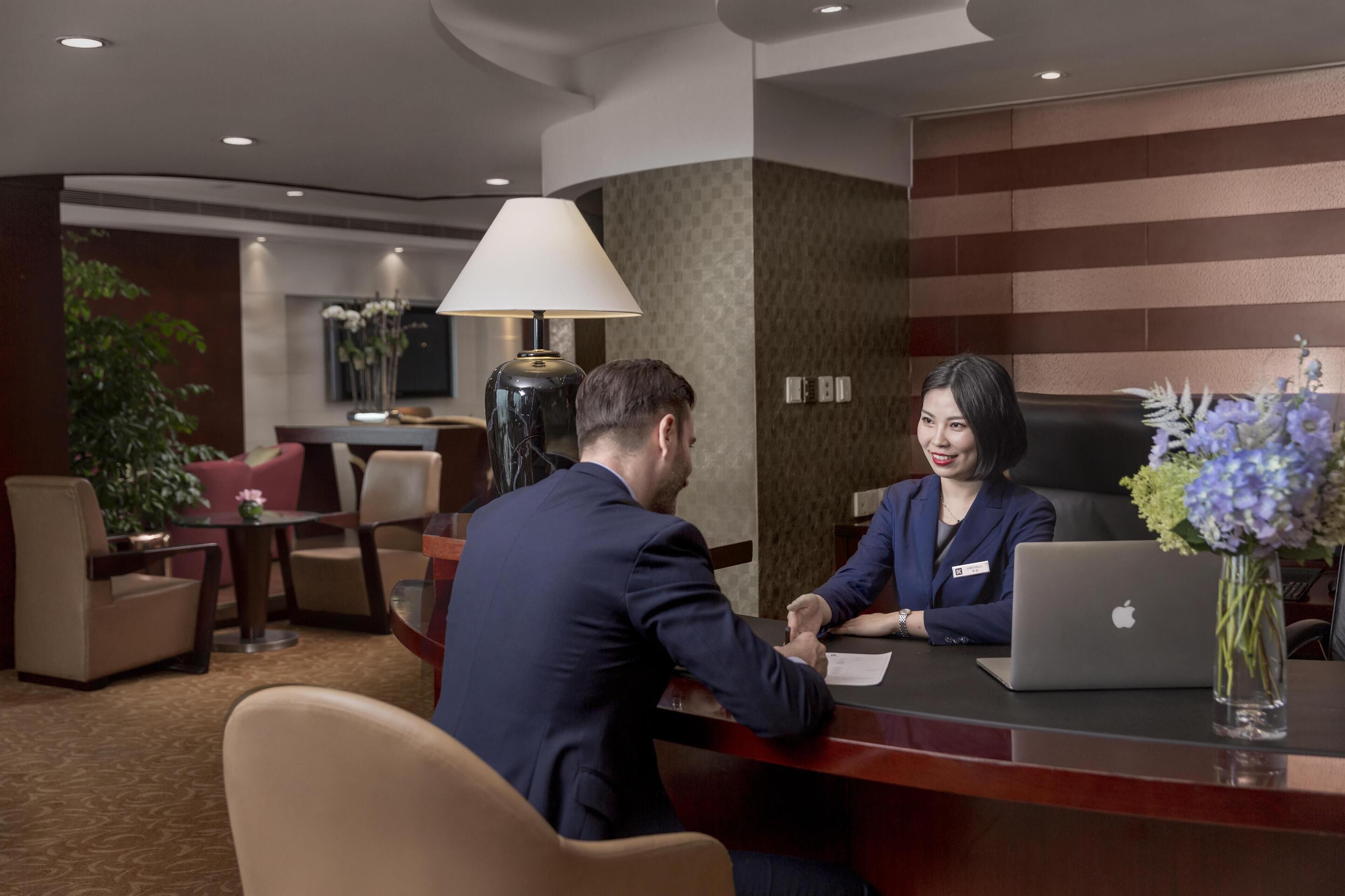 Een hotel receptiebalie met een man en een vrouw zittend, een laptop en een bloemenvaas.