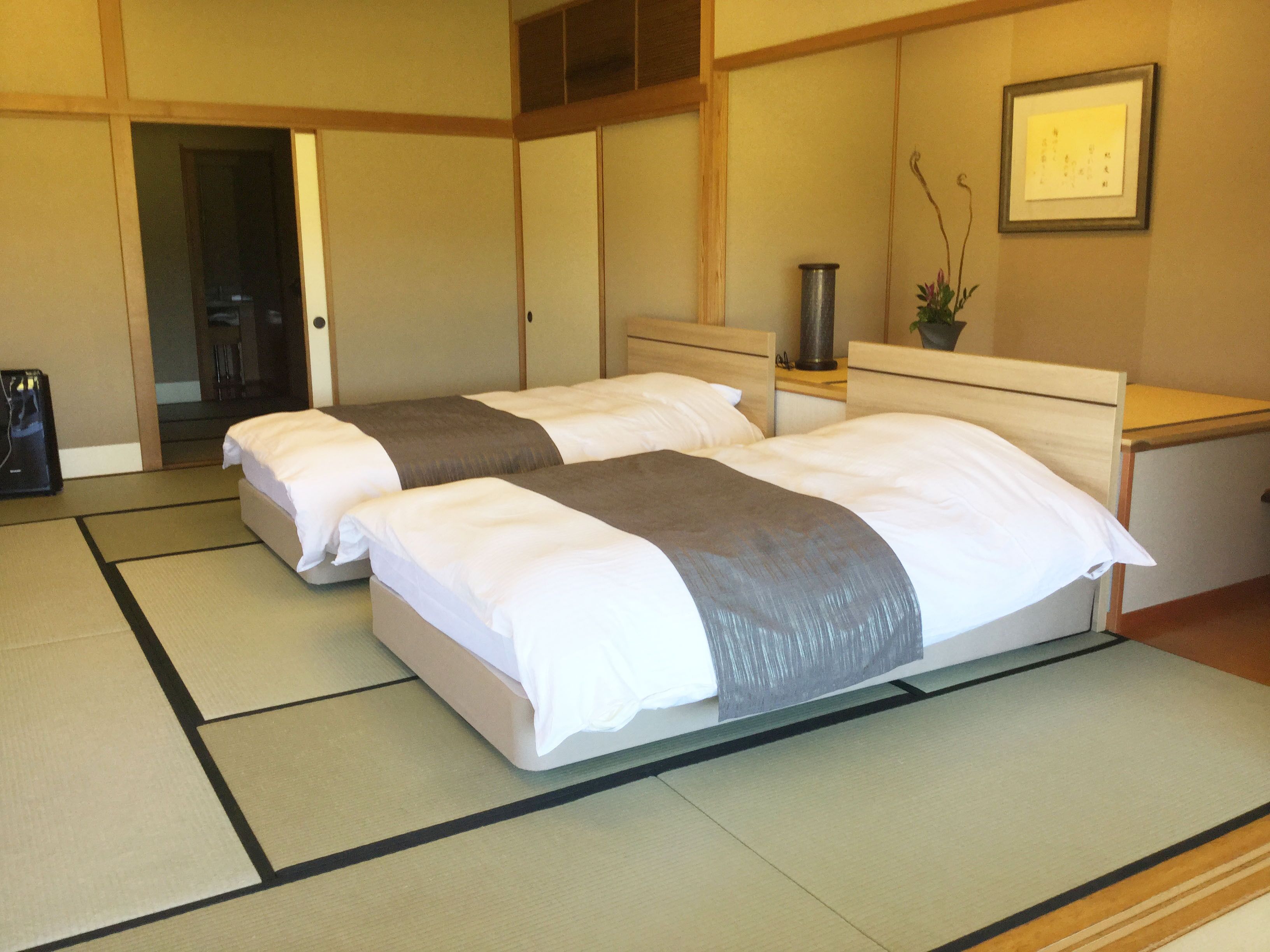 A hotel room with two beds, tatami mats, a bedside table with a lamp, and a framed picture on the wall.