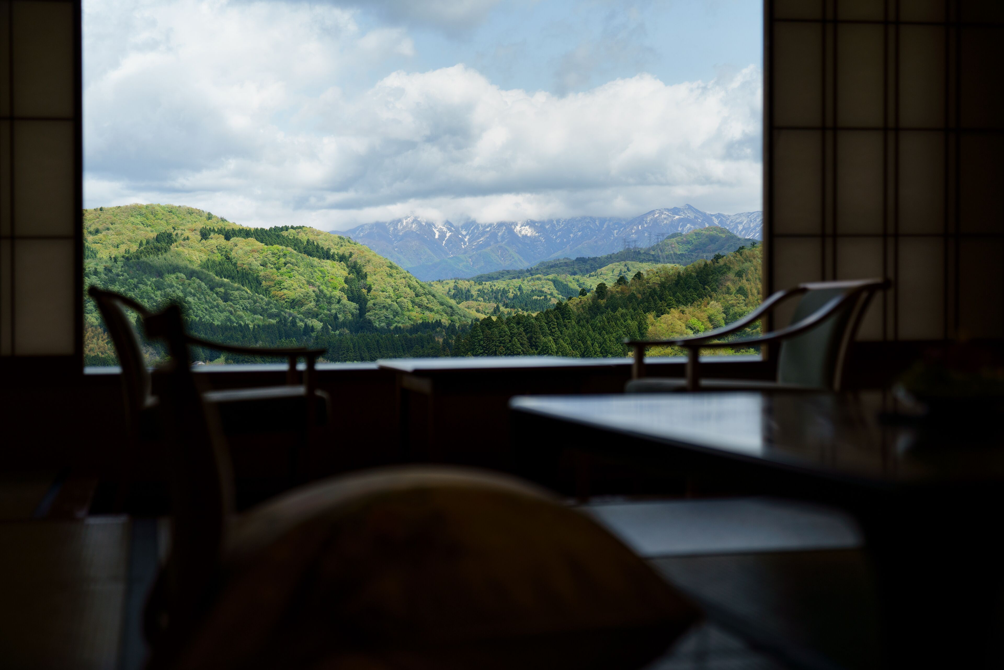 Ein Zimmer mit Blick auf eine Berglandschaft durch ein Fenster. Im Vordergrund stehen zwei Stühle und ein Tisch.