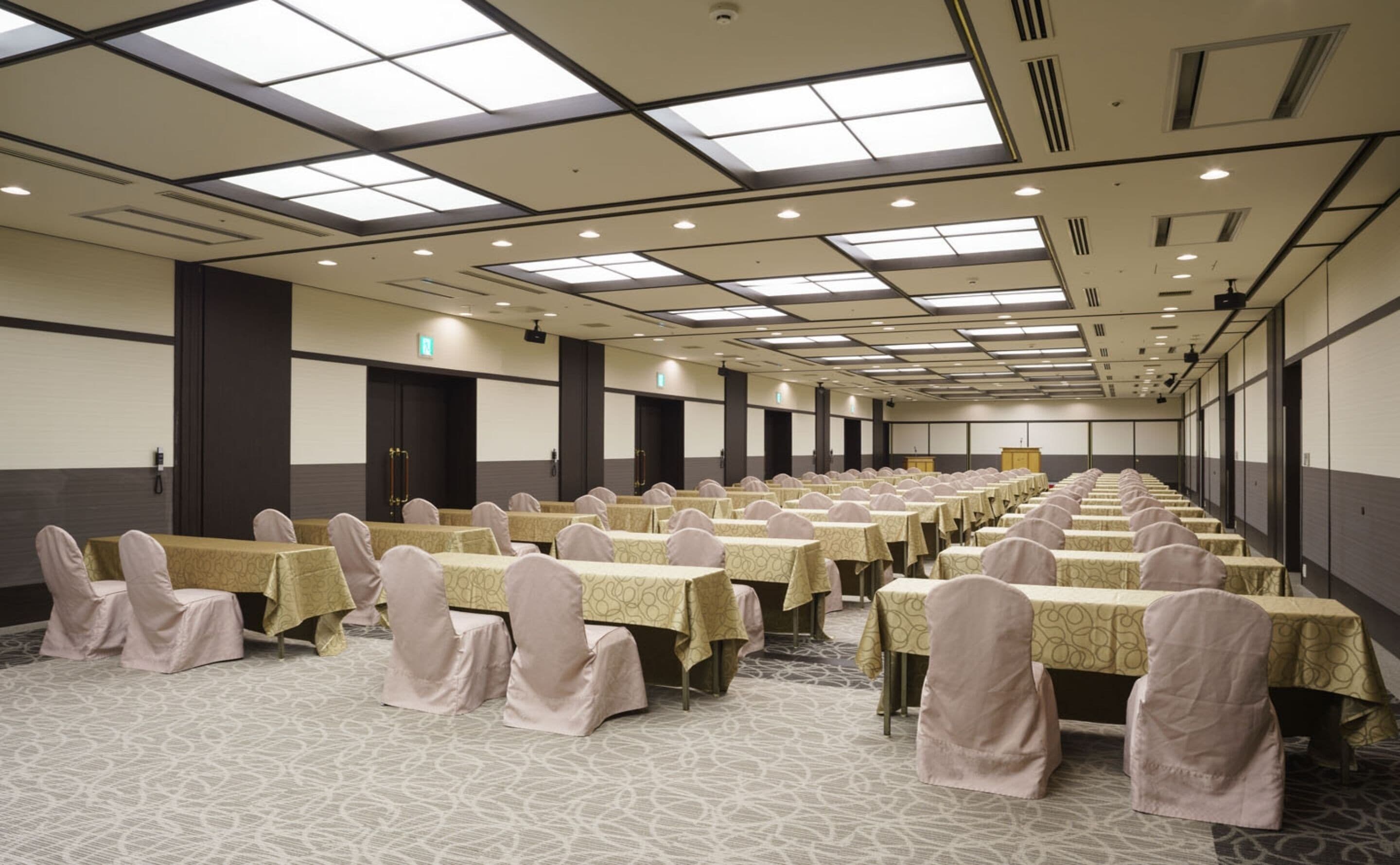 A conference room with rows of tables and chairs, large windows, and a high ceiling with recessed lighting.