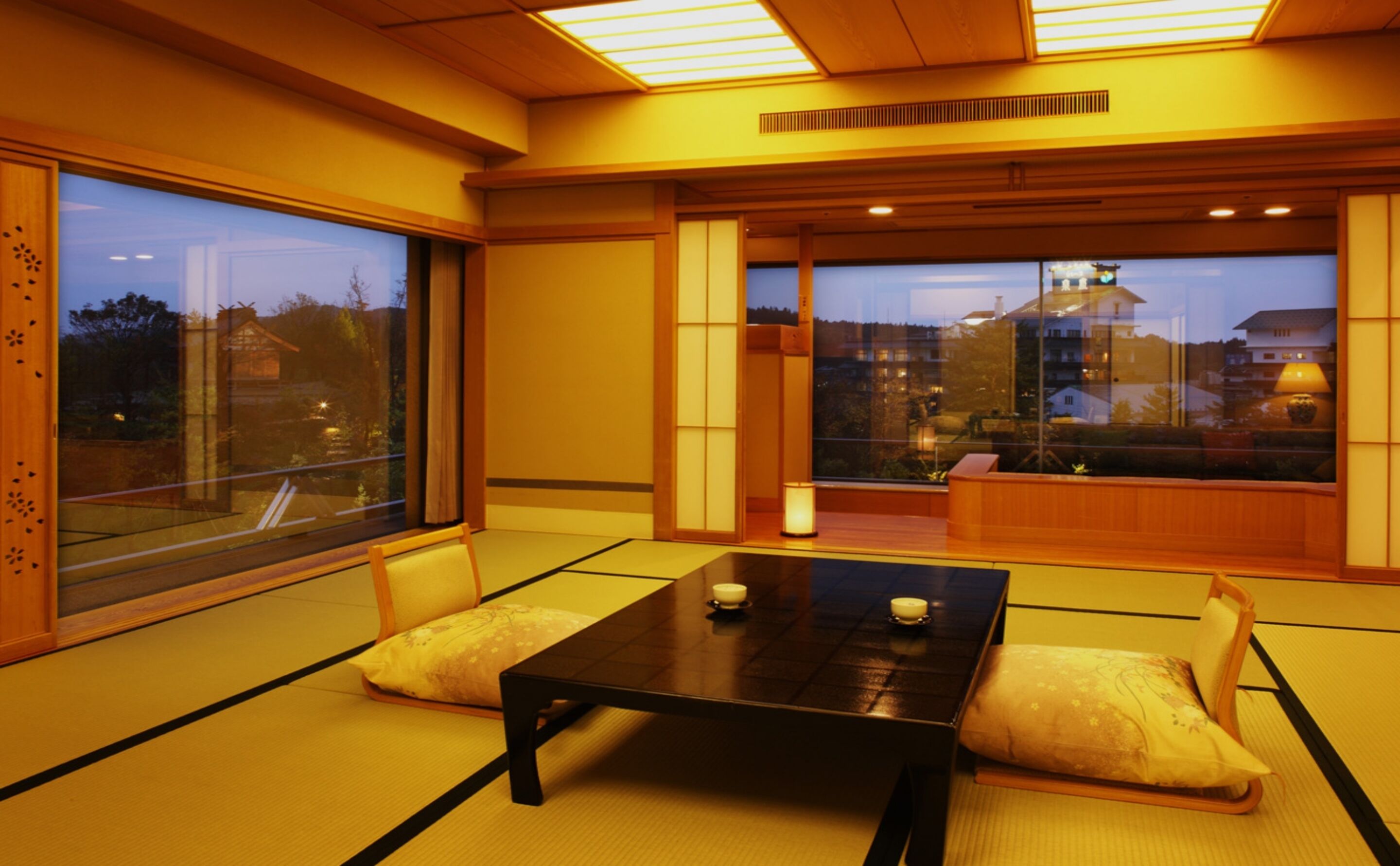 Ein traditioneller japanischer Raum mit Tatami-Boden, einem niedrigen Tisch, Kissen und großen Fenstern mit Blick nach außen.
