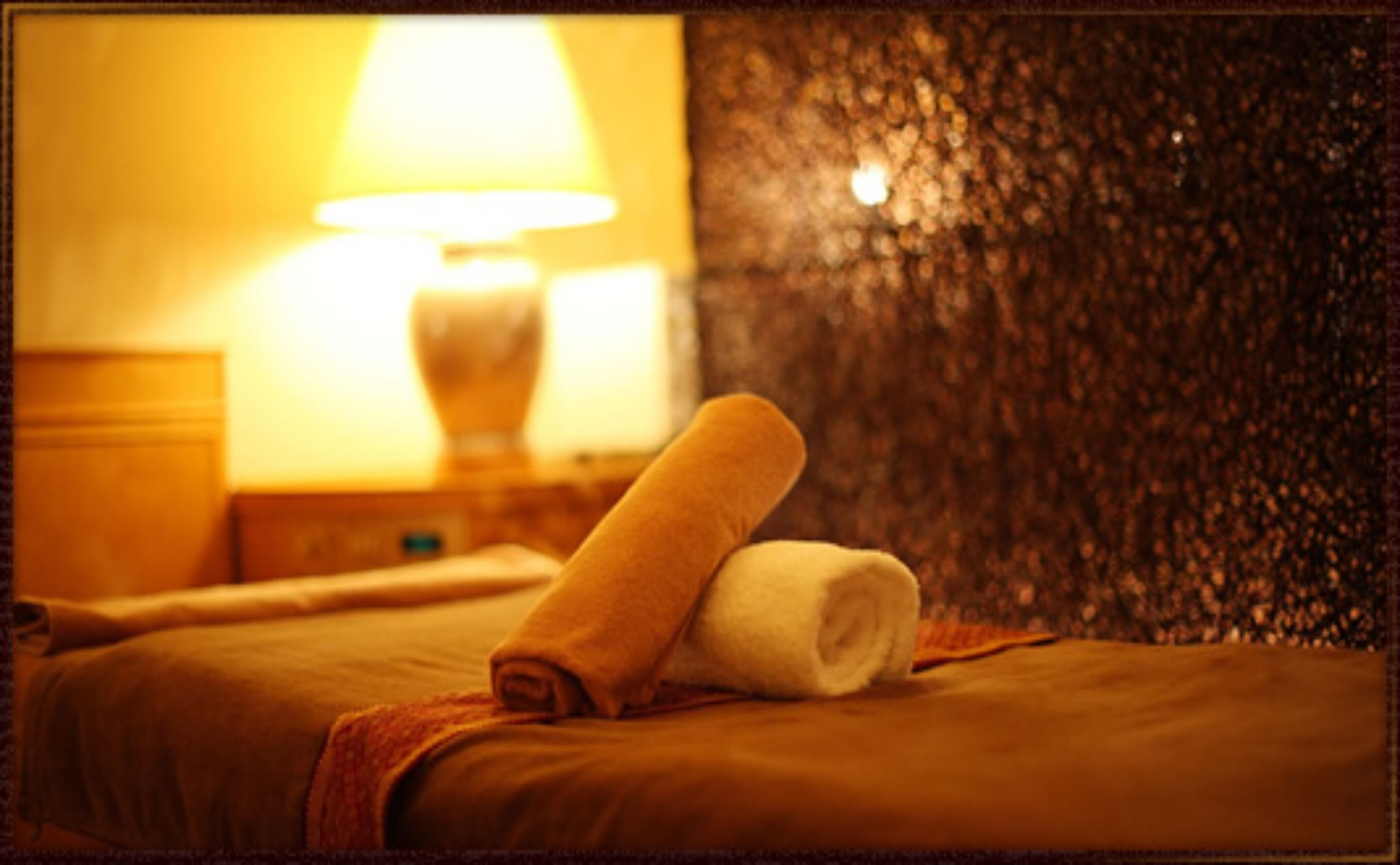 A hotel room with a bed, a rolled towel, and a lamp on a nightstand.