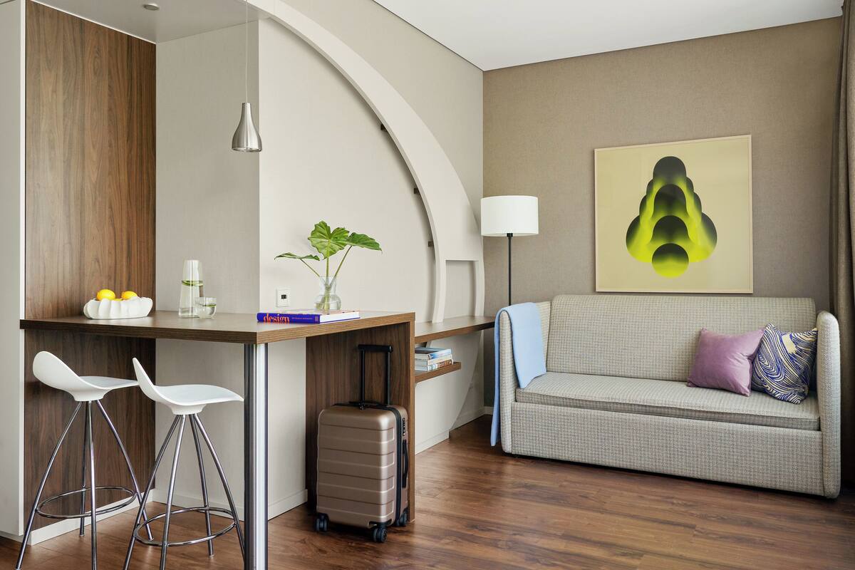 Une chambre d’hôtel moderne dotée d’une kitchenette, d’un canapé, d’un coin repas avec des tabourets et d’un grand tableau abstrait accroché au mur.