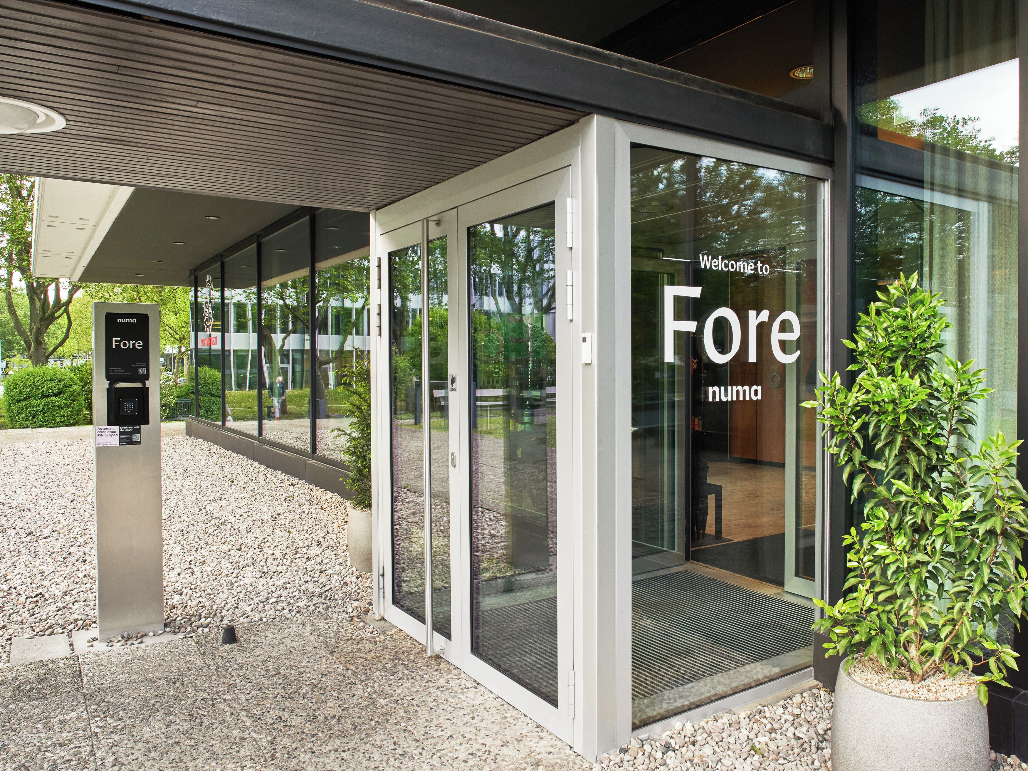Une entrée moderne avec une porte en verre, un kiosque de paiement numérique pour le stationnement et un pot de fleurs.