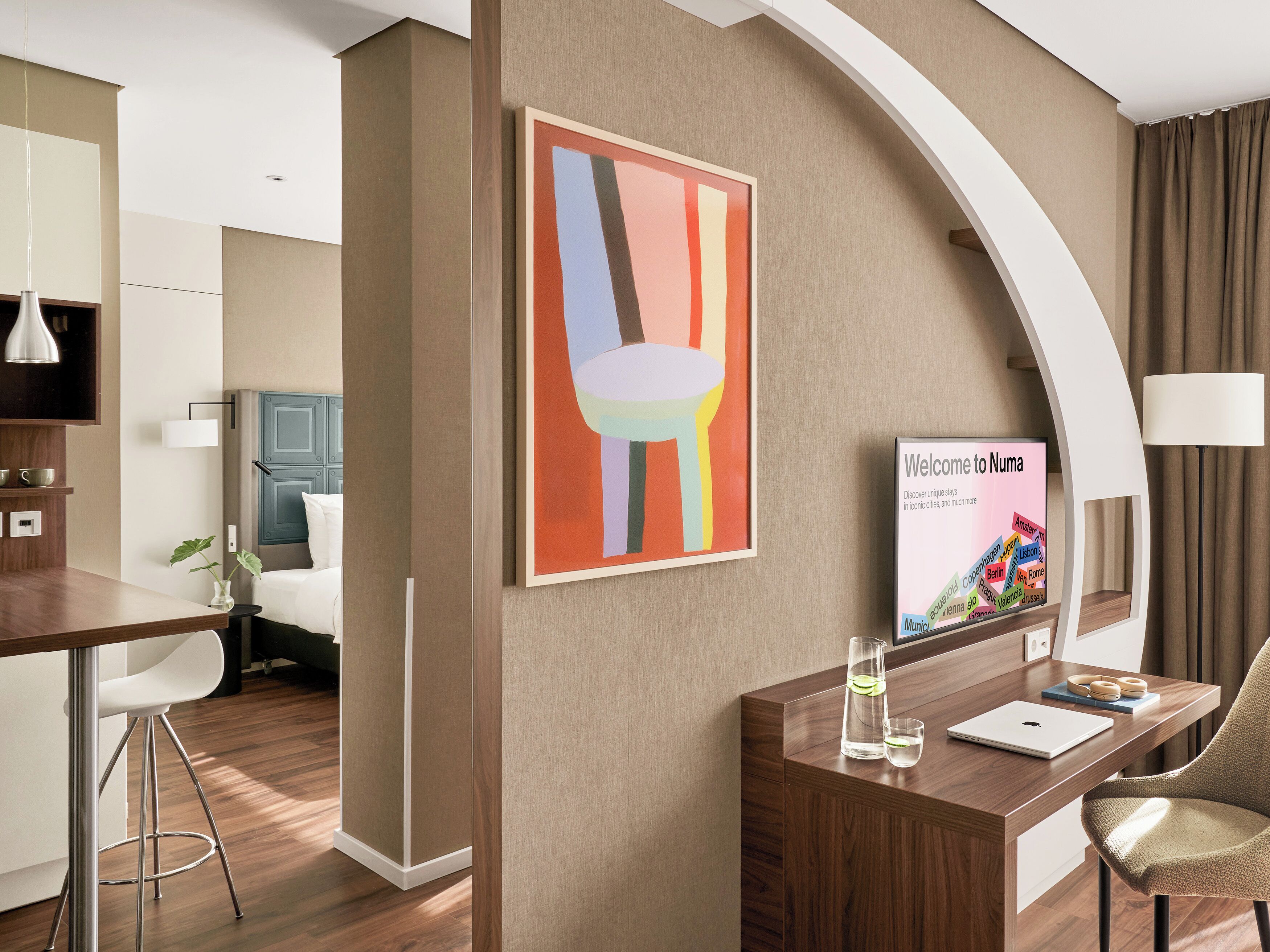 A modern hotel room with a wooden desk, a laptop, a glass pitcher, and a framed welcome sign.