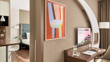 A modern hotel room with a wooden desk, a laptop, a glass pitcher, and a framed welcome sign.