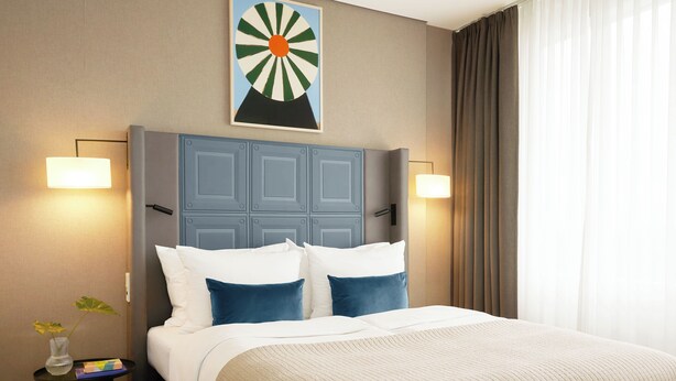 Une chambre d’hôtel avec un lit, une table de chevet, une lampe, un tableau et une fenêtre avec des rideaux.