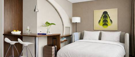 A modern hotel room with a bed, desk, two chairs, a lamp, a painting, and a suitcase.
