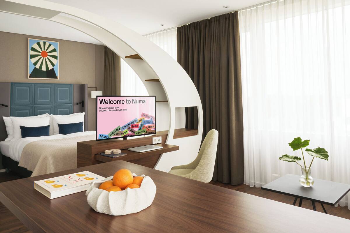 Une chambre d’hôtel moderne équipée d’un lit, d’un bureau avec un ordinateur portable, d’un téléviseur affichant le message « Bienvenue chez Numa », d’un vase avec des plantes et d’un saladier rempli d’oranges.