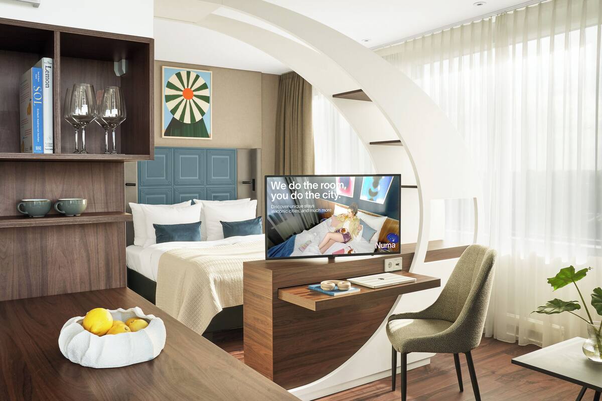 Une chambre d’hôtel moderne équipée d’un lit, d’un bureau, d’une télévision et d’un coin repas agrémenté d’un panier de fruits.