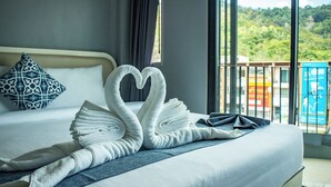 A hotel room with a bed, a heart-shaped towel arrangement, a patterned pillow, and a balcony with a view of greenery.