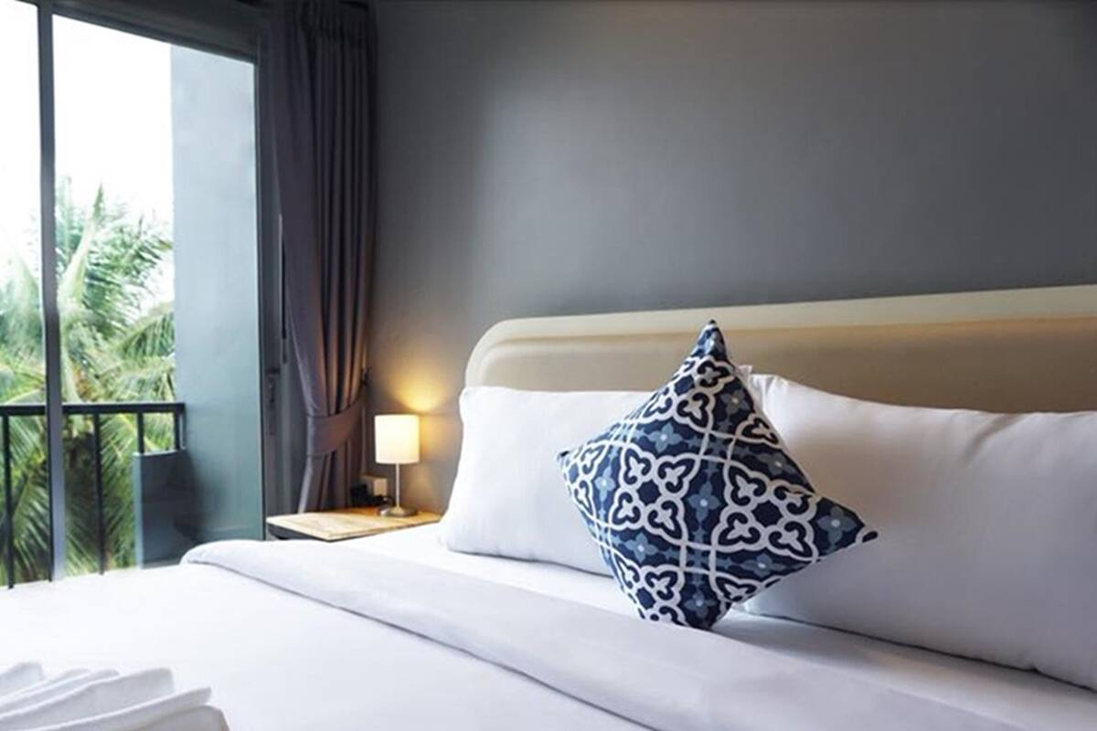 Une chambre d’hôtel avec un lit, une table de chevet avec une lampe et une vue sur les palmiers depuis un balcon.