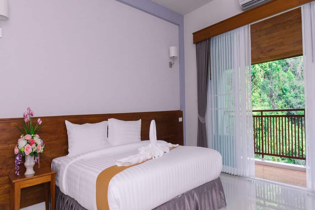 Une chambre d’hôtel avec un grand lit, une tête de lit en bois, une table de chevet ornée d’un vase de fleurs et un balcon donnant sur un paysage verdoyant.