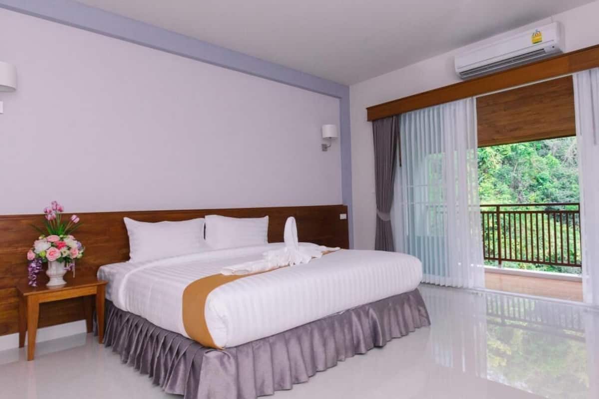 Une chambre d’hôtel avec un grand lit, une table de chevet ornée d’un vase de fleurs, une tête de lit en bois, un balcon donnant sur un paysage verdoyant et un appareil de climatisation.