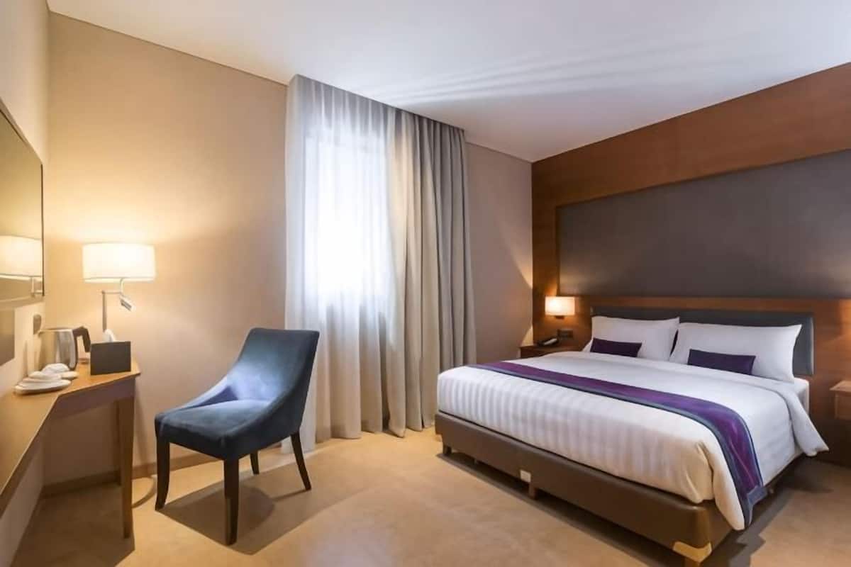 A hotel room with a bed, a desk, a chair, a nightstand, and a window with curtains.