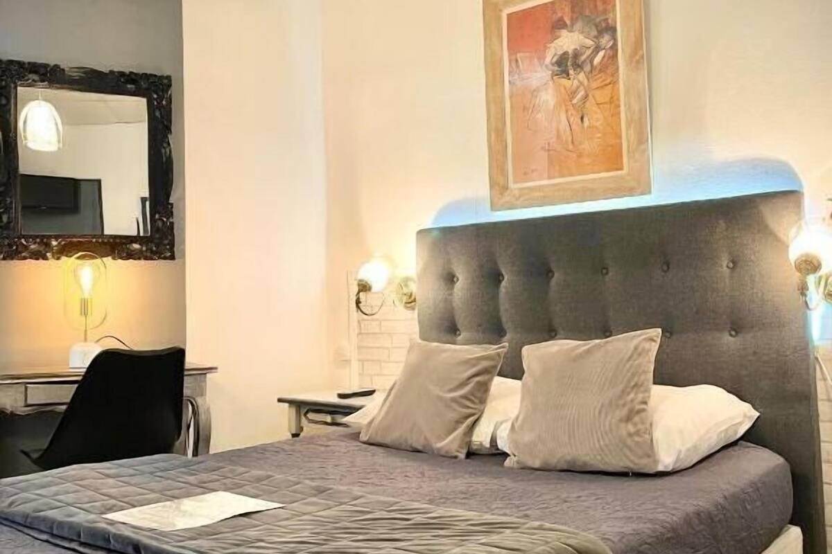 Quarto com cabeceira estofada, cama com roupa de cama cinzenta, mesa de cabeceira, cadeira, escrivaninha com luminária e um quadro na parede.