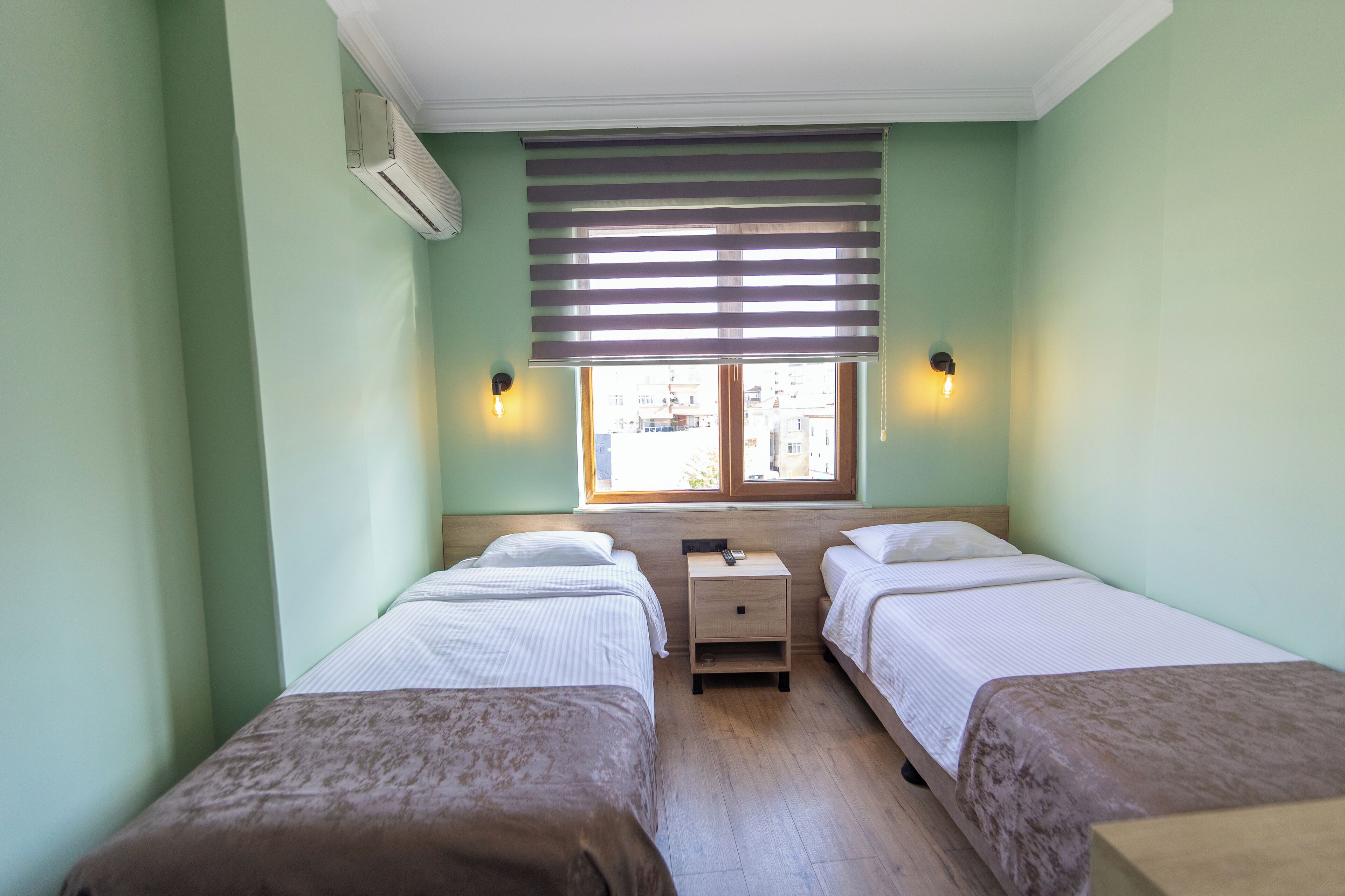 A room with two beds, a nightstand, a window with blinds, and wall-mounted lights.