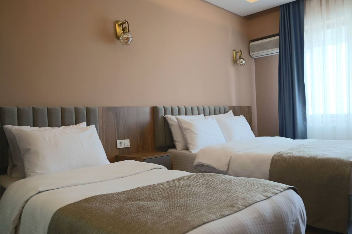 A hotel room with two beds, a wooden headboard, a wall-mounted air conditioner, and a window with curtains.