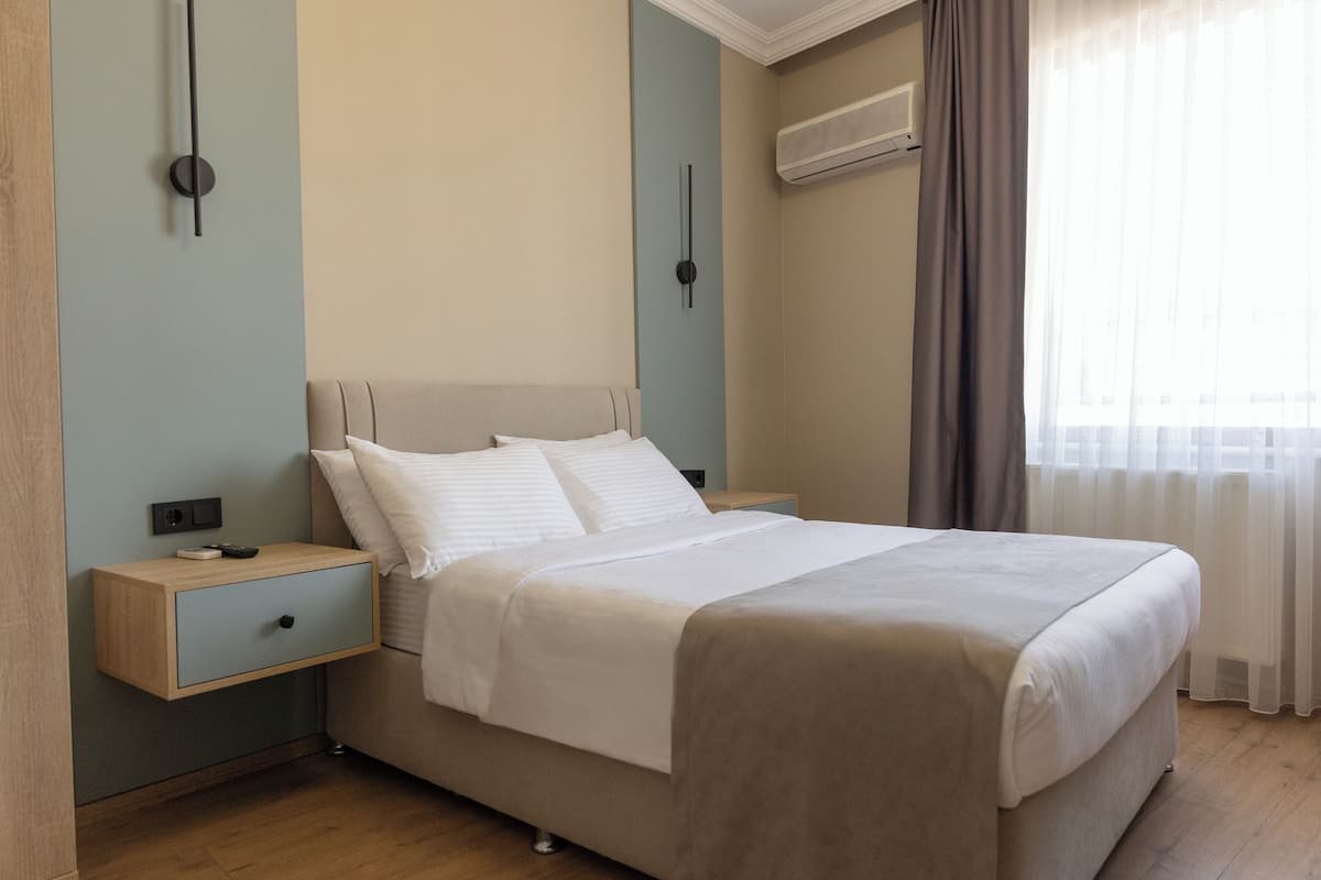 A neatly made bed with white linens, a wooden nightstand with a drawer, and a wall-mounted air conditioning unit.