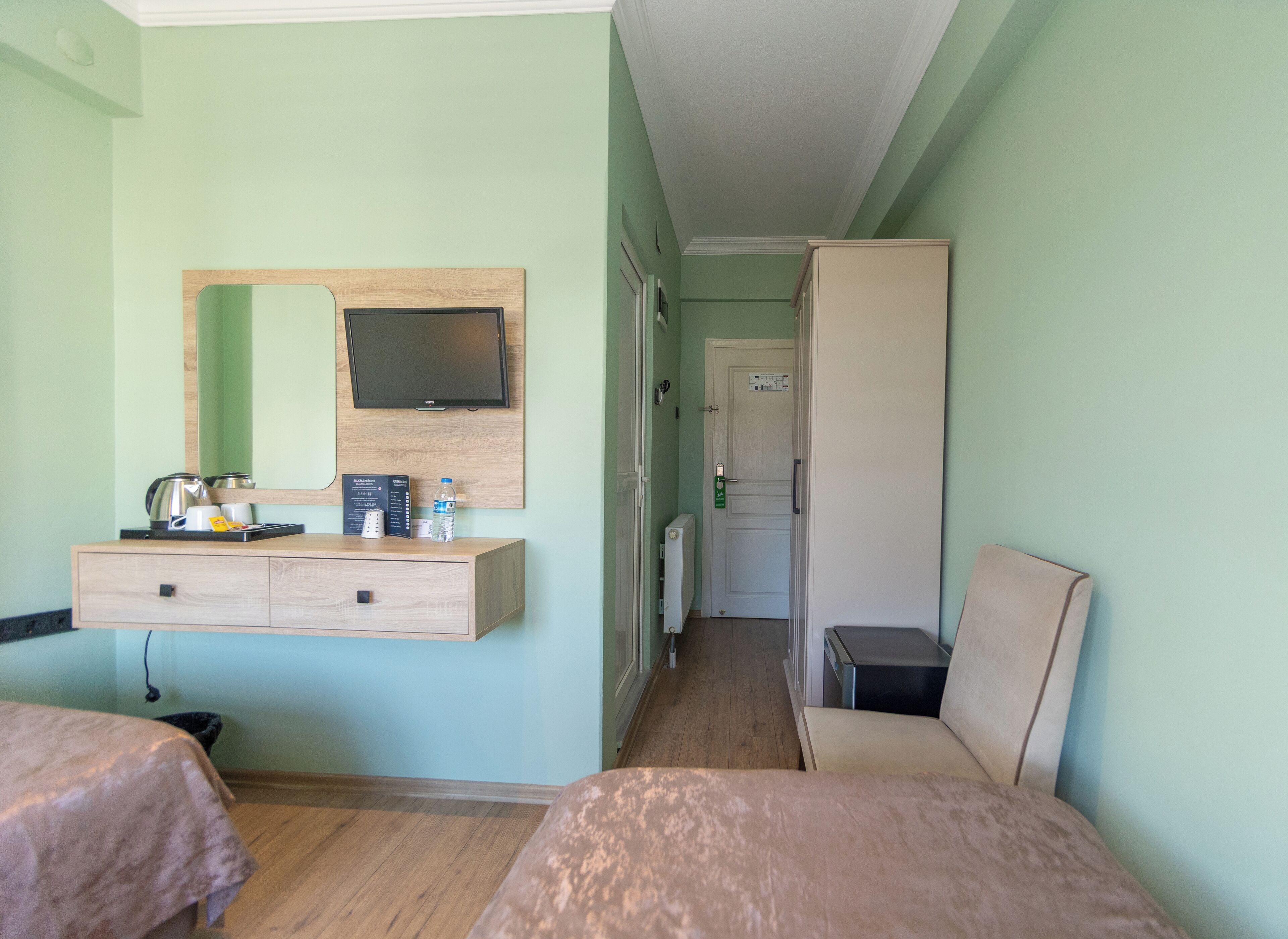 Quarto de hotel com cama, mesa de cabeceira com chaleira, espelho, televisão e guarda-roupa.