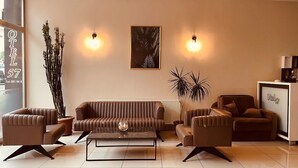 Un salon moderne avec un canapé en cuir marron, deux fauteuils, une table basse et un buffet. On y trouve deux lampes fixées au mur, une œuvre d’art encadrée et une plante en pot.