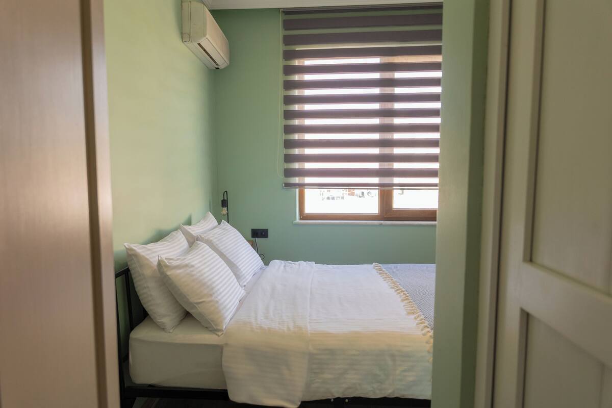 A room with a bed, a window with blinds, and an air conditioning unit on the wall.