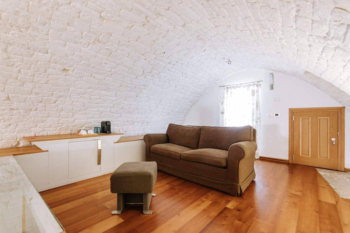 Een loft-achtig vertrek met een witte baksteen plafond, een bruine bank, een houten salontafel en een houten deur.