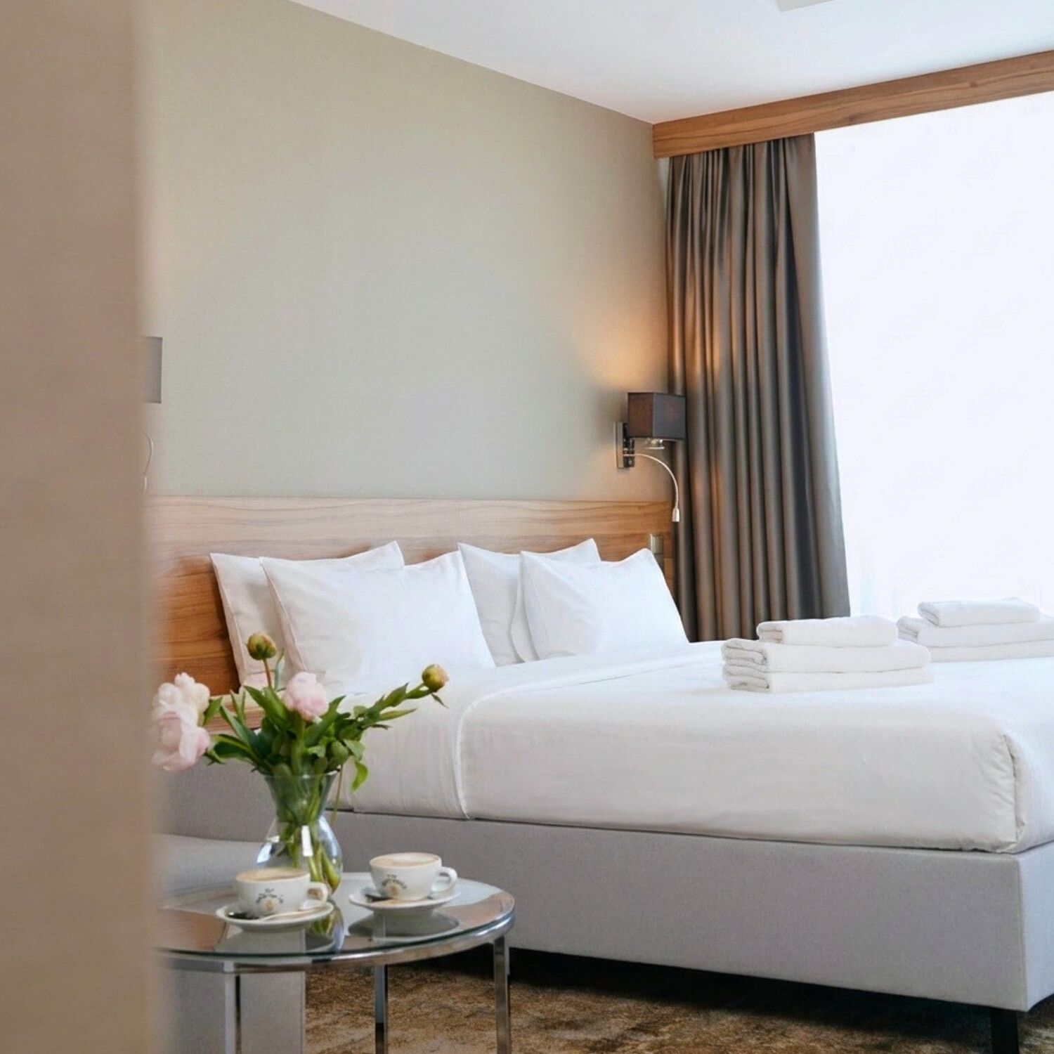 Ein Hotelzimmer mit einem Bett, einem Nachttisch mit Blumenvase, einer Tasse und einem Untertasse sowie einem Fenster mit Vorhängen.