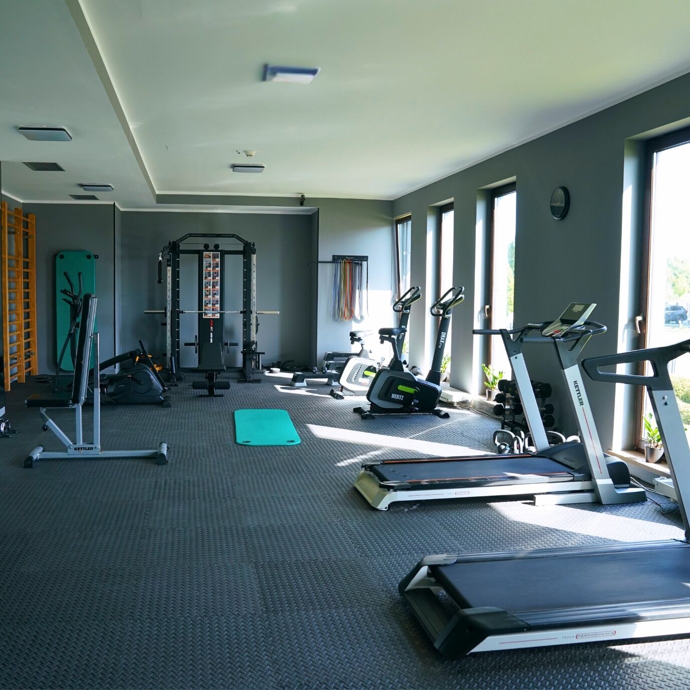 Un gimnasio con equipo de ejercicio, un estante fijado a la pared y grandes ventanales.