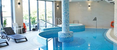 An indoor swimming pool with a tiled floor, a central column, and a sunlit area with lounge chairs.