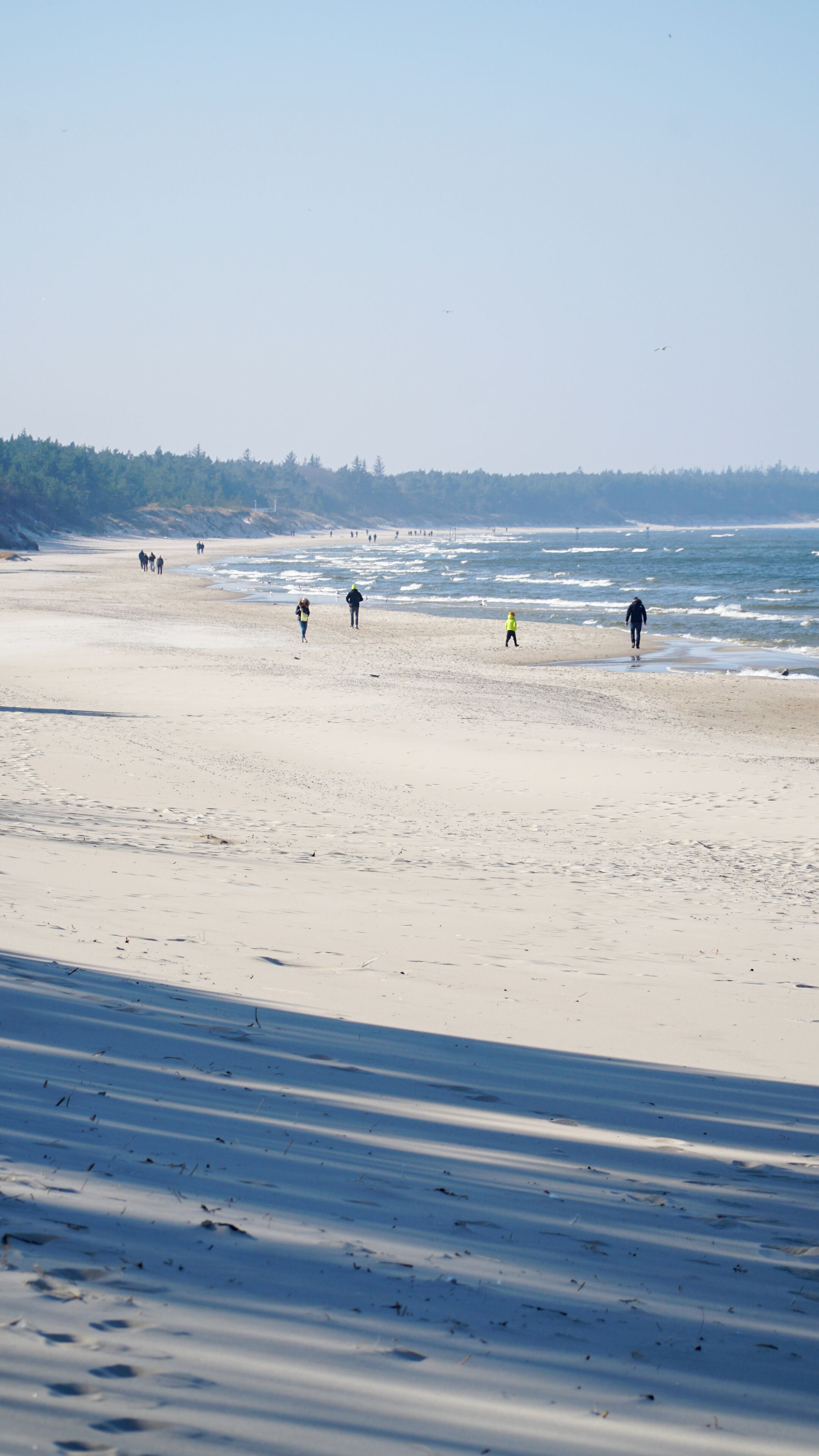 Una playa con gente caminando por la orilla, cielo despejado y un área boscosa al fondo.